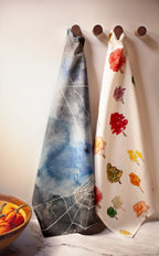 Two decorative towels with spider web and leaf patterns hanging on wooden hooks against a white wall.