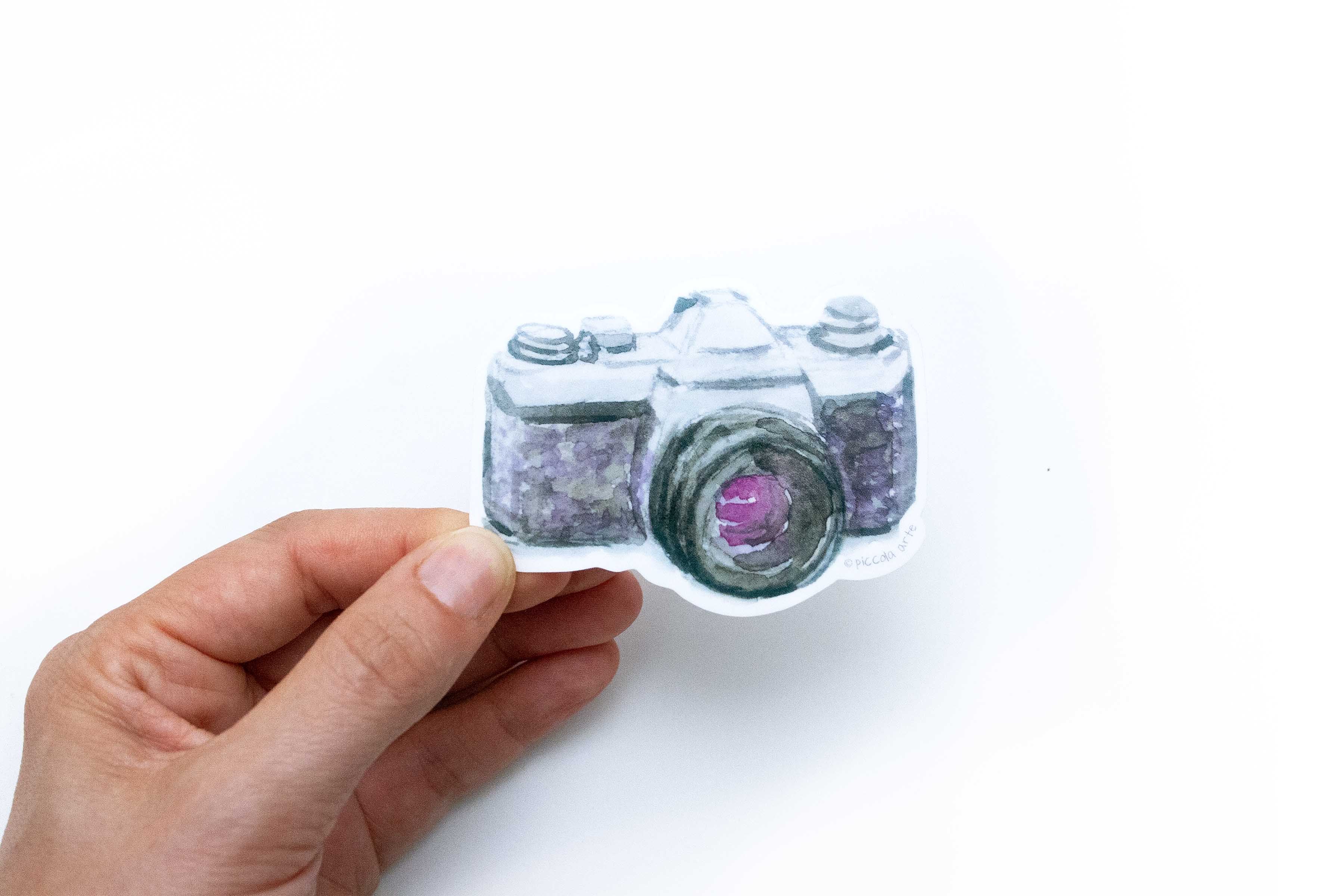 Hand holding a camera-shaped sticker on a white background