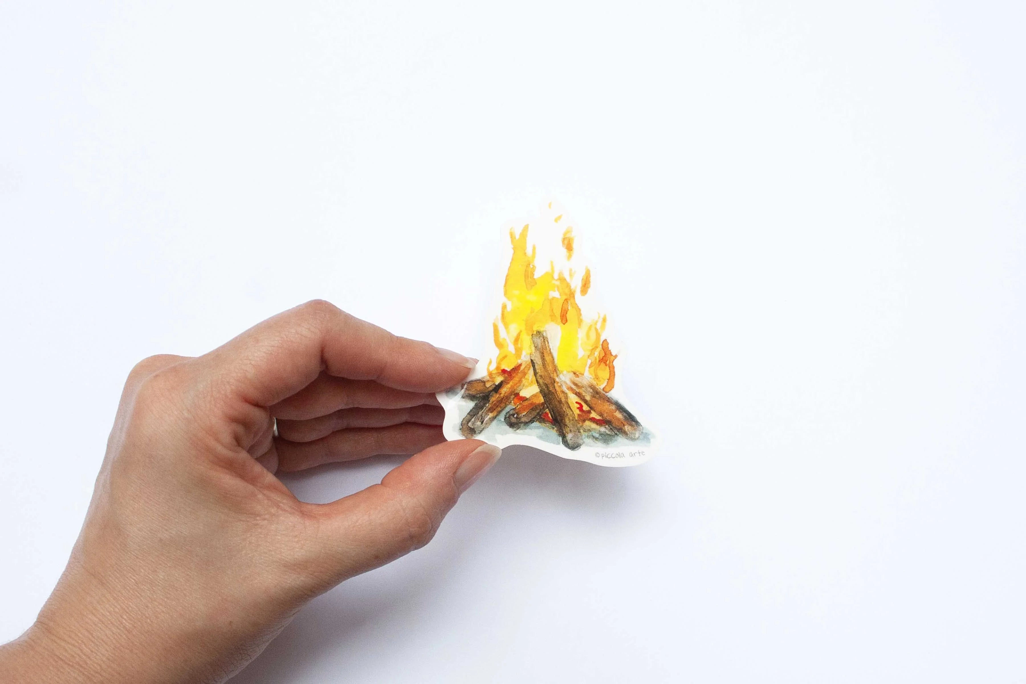 Hand holding a sticker cutout of a campfire with flames on a white background.