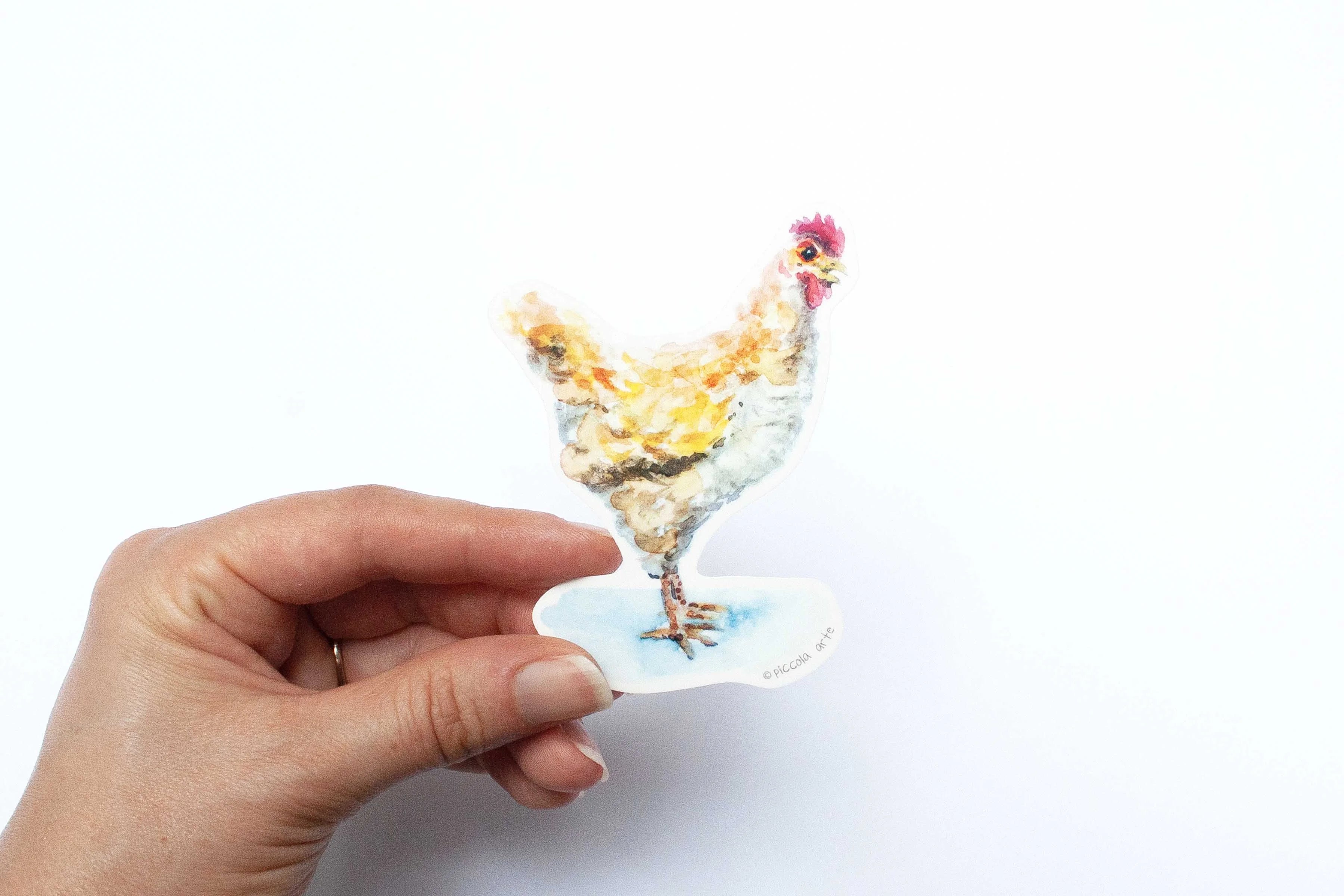 Hand holding a small sticker chicken figurine on a white background.