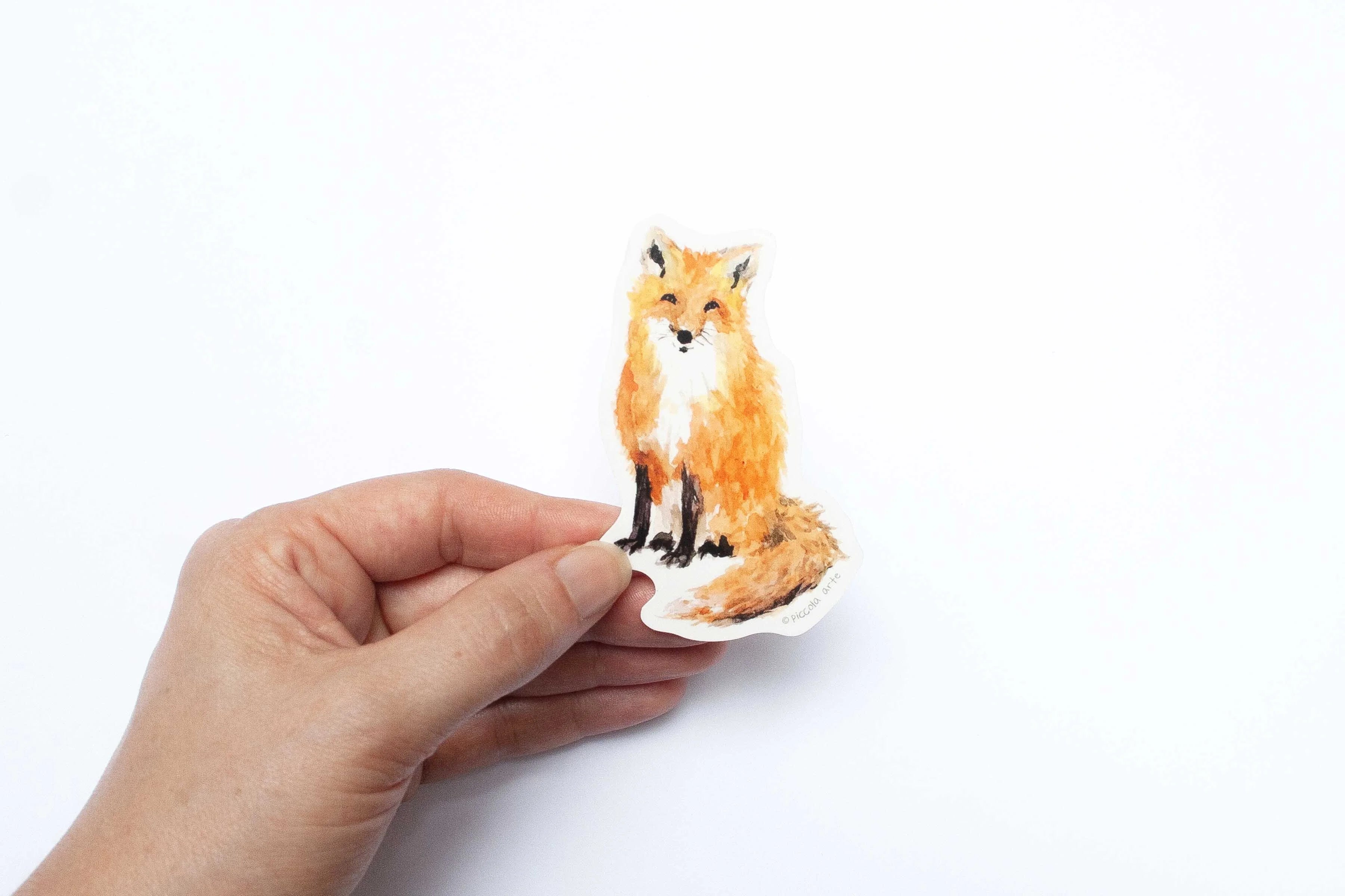 Hand holding a small fox sticker on a white background.