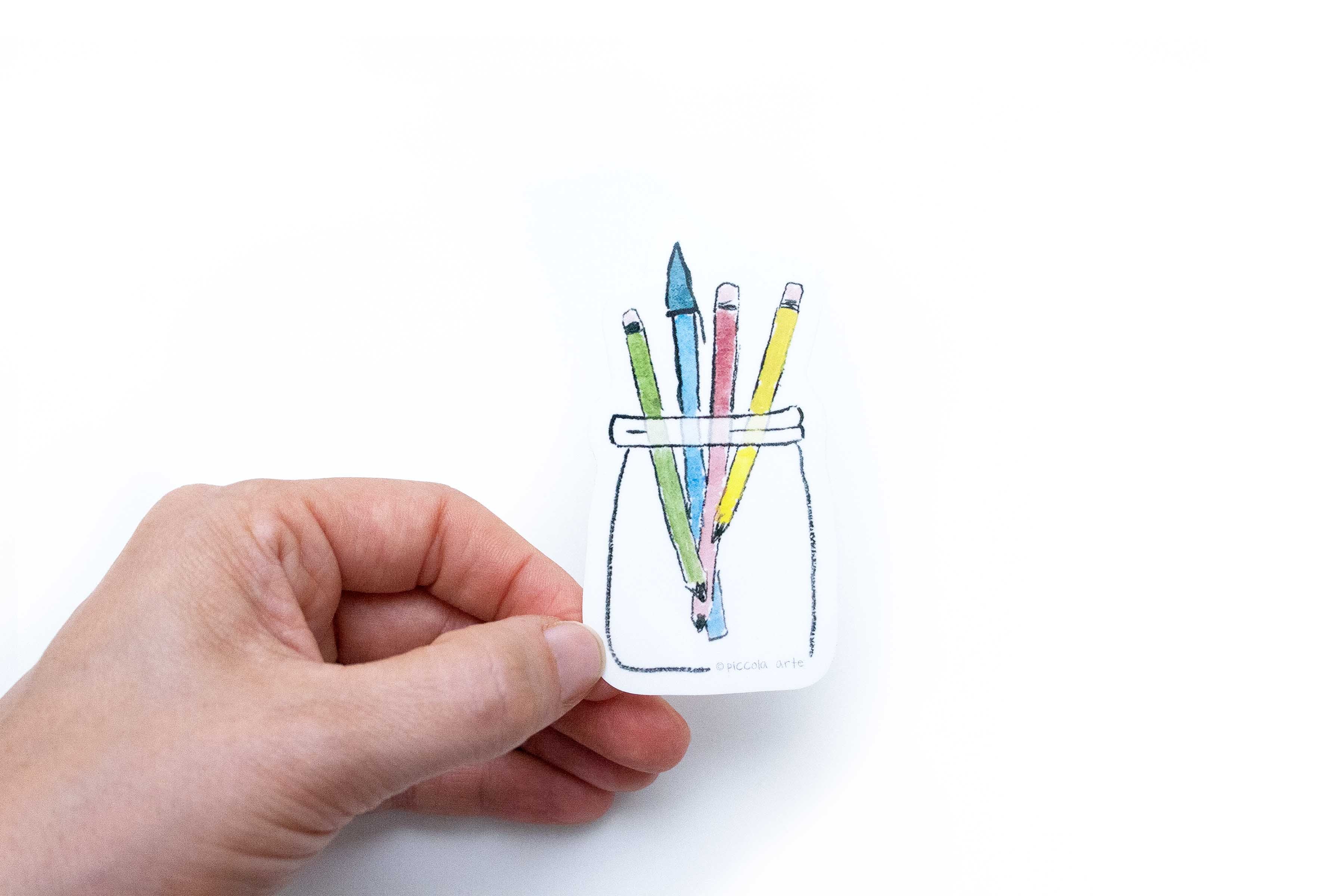 Hand holding a small jar with colorful pencils against a white background