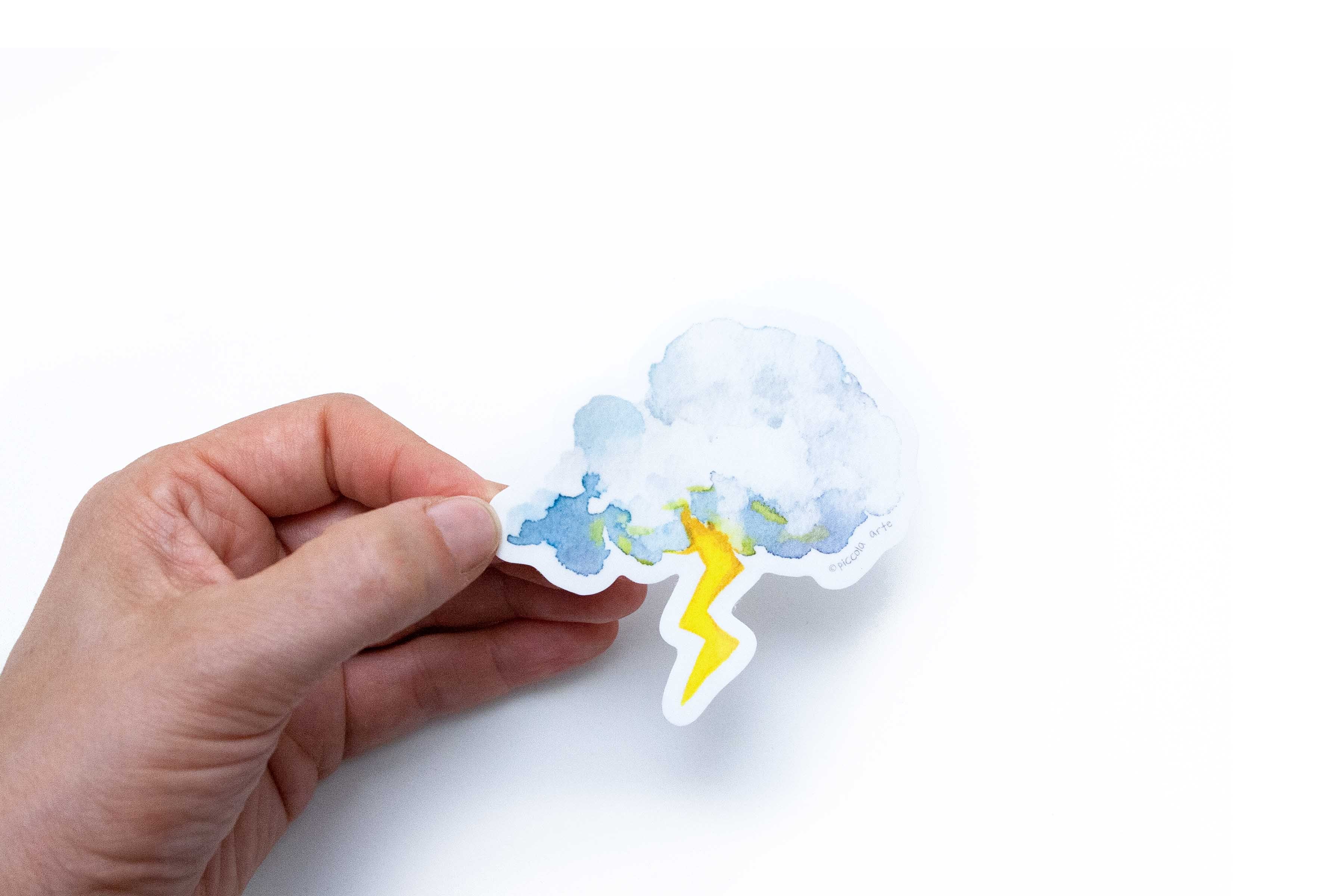 Hand holding a weather-themed sticker with a cloud and lightning bolt on a white background