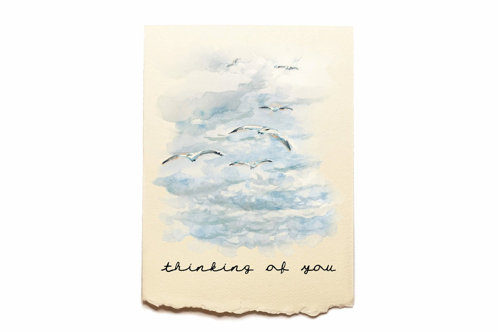 Card with watercolor illustration of birds flying in a cloudy sky and 'thinking of you' text on a beige background.