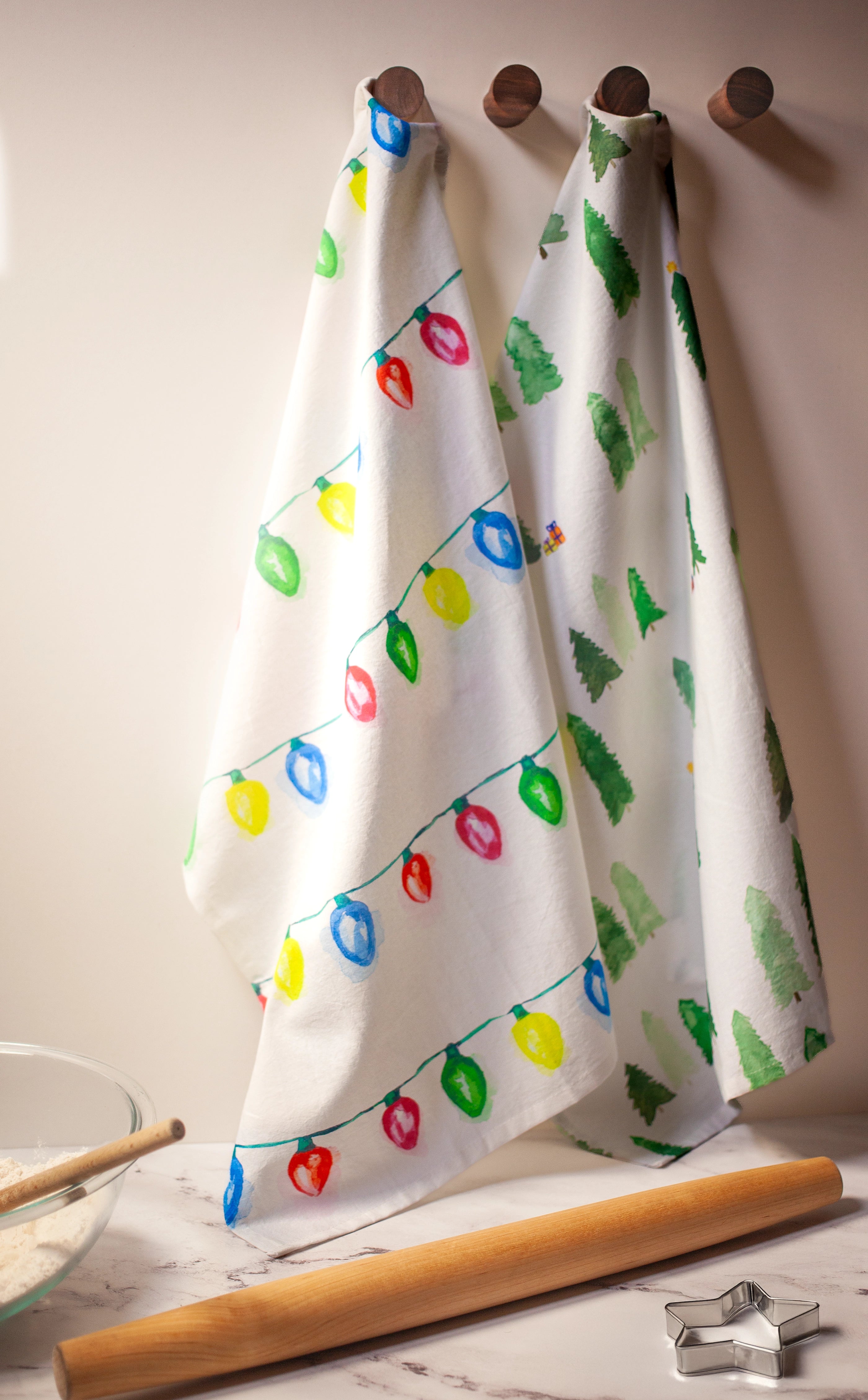 Two kitchen towels with Christmas-themed designs hanging on hooks, with a rolling pin and cookie cutter on a countertop.